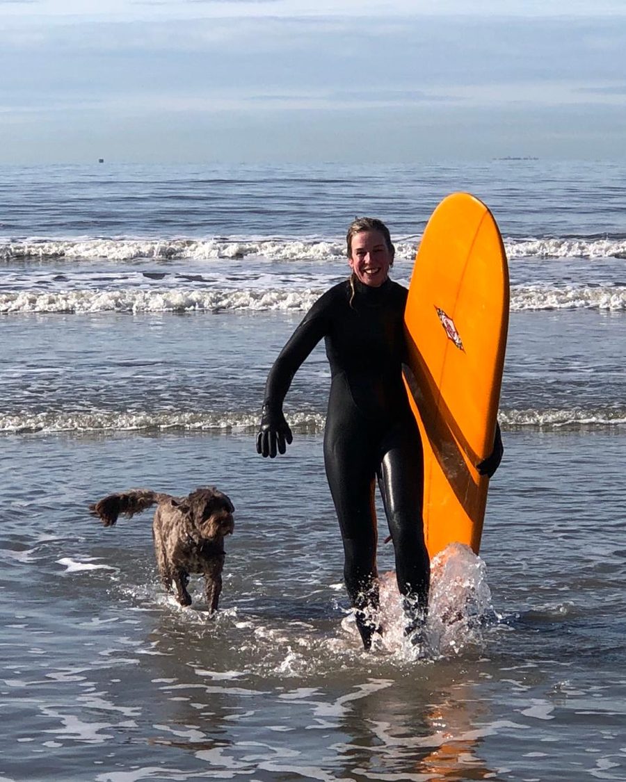 Mandy walking out of the sea with her longboard and her dog